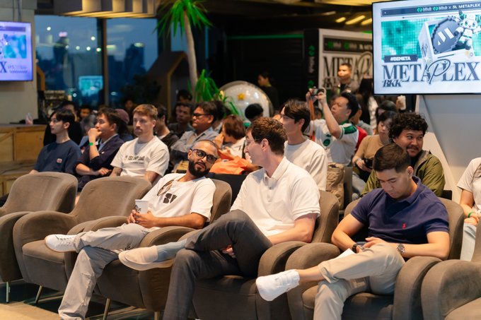 A group of casually dressed people sitting in armchairs at what appears to be a METAPLEX event, watching a presentation with screens visible in the background.