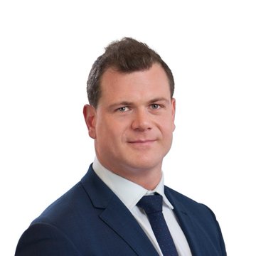 A professional headshot shows a man with short brown hair wearing a navy blue suit, white dress shirt, and dark tie, smiling slightly against a plain white background. The image accompanies a news quote attributed to IREN co-founder Daniel Roberts, highlighting the company's strong demand for compute capacity and significant progress toward its 2026 revenue goals.