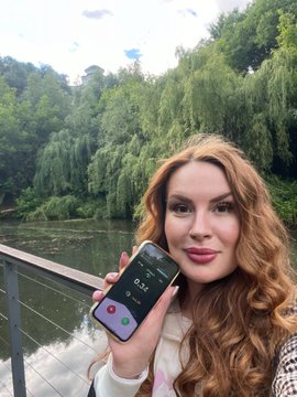 The image features a woman holding up her cell phone, which has a photo of herself taken at a park or lake. The background includes trees and water, creating a serene atmosphere for the photograph. The caption provided seems unrelated to the main subject matter but is still present in the image.