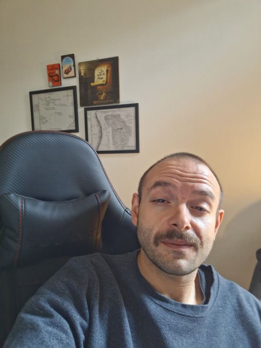 The image features a man sitting in an office chair, wearing a blue sweater with his eyes closed as if he's meditating or taking a break from work. The background includes several framed pictures hanging on the wall above him.