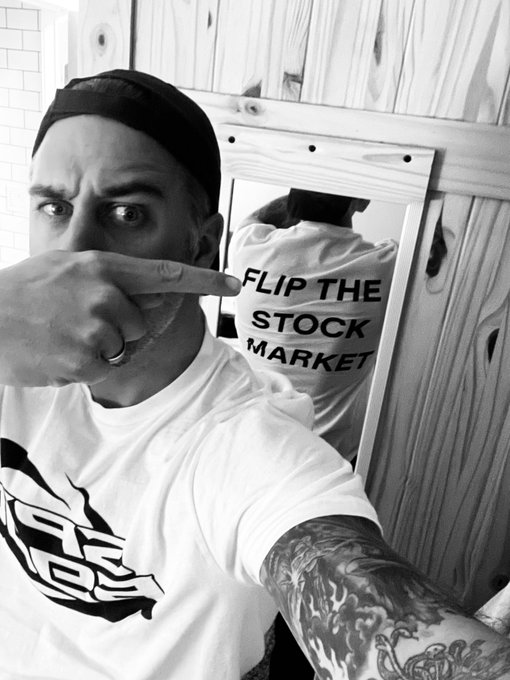 A black-and-white selfie of a tattooed man wearing a baseball cap backwards and a t-shirt, pointing at a mirror reflection that reveals text reading "FLIP THE STOCK MARKET" on his shirt.