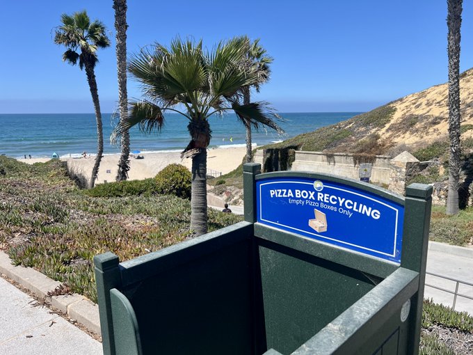 A photograph of a green pizza box recycling bin labeled 'Empty Pizza Boxes Only' positioned on a coastal pathway overlooking a sandy beach, ocean, and palm trees.