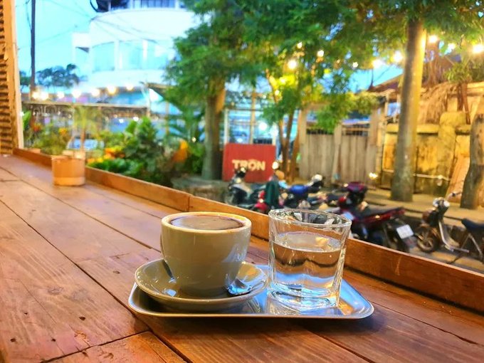 A coffee cup and a glass of water sit on a metal tray atop a wooden counter, with motorcycles parked in the background and a red sign partially reading 'TRÒN'.