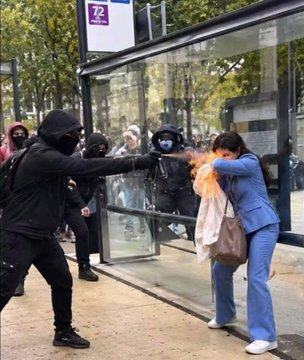 A chaotic confrontation at a bus stop shows masked individuals in black extending a lit flare or fire toward a woman in a blue suit who is recoiling and shielding her face.