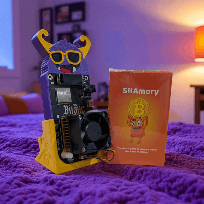 The image features a purple bedspread with an electronic device, including a robot toy and a book titled "SHAmory" that appears to be about mining. The robot toy has a pair of sunglasses and a hat, while the book is open on the bedspread, possibly discussing the concept of mining or its applications in real life. The image seems to be related to the post because it contains text that reads: "Real mining beats screen lectures. SHAmory Satoshi Bitaxe combo for kids! Learn all about mining while seeing it in action!"