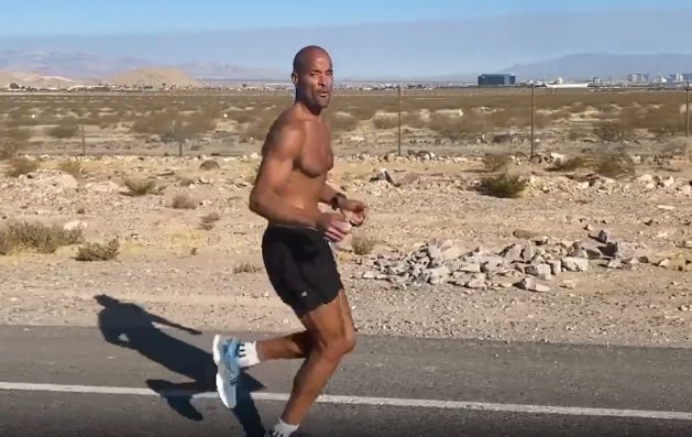 A shirtless, muscular Black man runs along a paved road through a desert landscape, wearing black shorts and white running shoes with a mountainous horizon in the background.