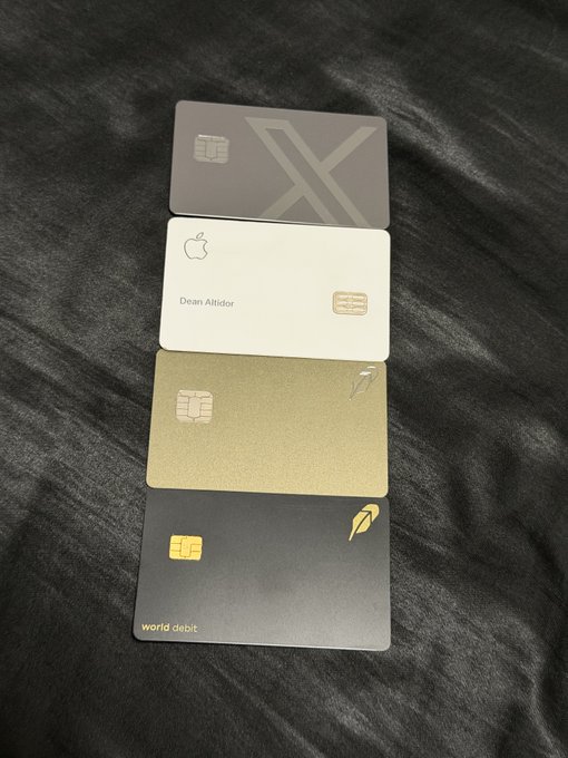 Four credit cards are arranged in a vertical stack on a black textured surface. The cards include a gray card with an 'X' logo, a white Apple Card issued to 'Dean Altidor', a metallic gold card, and a dark card labeled 'world debit'.