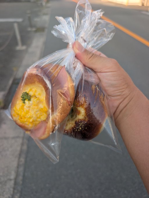 A hand holds up a clear plastic bag containing two pieces of bread, one with corn and parsley filling and another with what appears to be a meat or chocolate filling, against a street background.