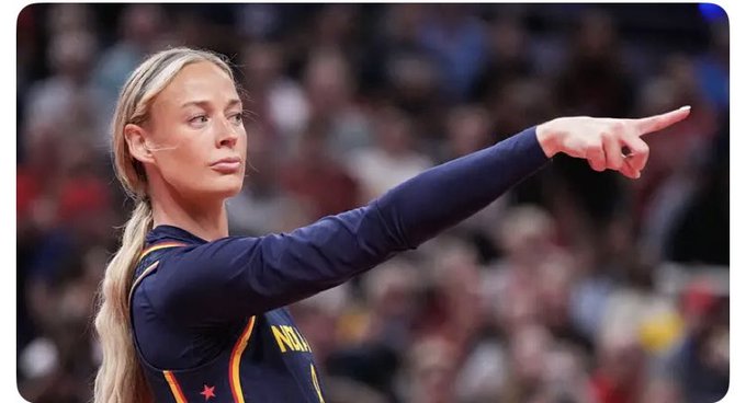 A blonde female athlete wearing a navy long-sleeve jersey, pointing her finger toward the right side of the frame, with a blurred crowd in the background.