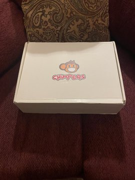 A white box with a red 'HAPPYERS' logo featuring a cartoon face sits on a maroon cushion next to a patterned pillow.