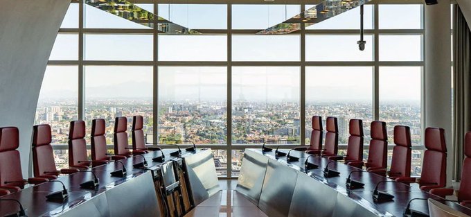 A formal conference room with a long table, red leather chairs, and panoramic windows overlooking a city skyline, representing the high-rise boardroom setting requested for a live stream of Ryan Myher taking his official seat following Ginkgo Bioworks ($GStock) board appointment.