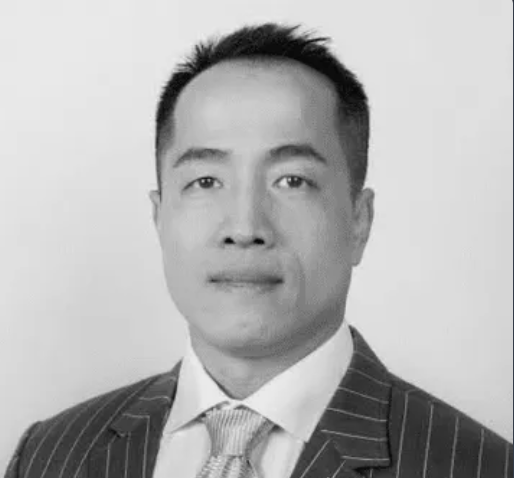 A black-and-white professional headshot of an Asian man wearing a pinstriped suit and tie, looking directly at the camera with a neutral expression.