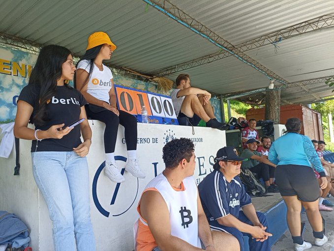 A photo of spectators sitting in a shaded grandstand area, including two women wearing 'berlin' branded shirts, and a man wearing a white tank top with the Bitcoin '₿' logo printed on it.