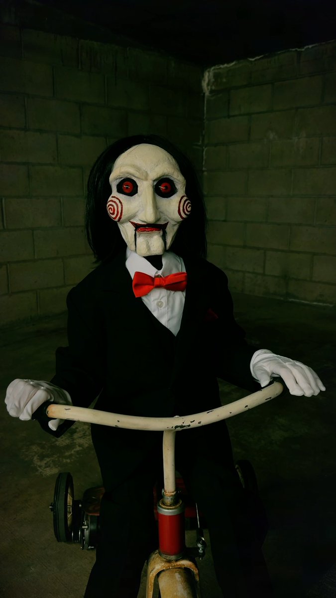 The "Billy the Puppet" figure from the *Saw* film franchise, wearing his iconic black suit, white shirt, red bow tie, and grotesque mask, rides a child's tricycle in a dark concrete room — a horror-themed visual evoking the chilling, game-like atmosphere referenced in the accompanying "Pond0x.com / The game continues." post.