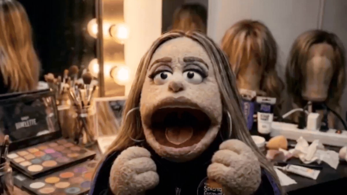 The image features a woman with blonde hair wearing a black shirt, sitting at a vanity table surrounded by various beauty products such as makeup brushes, lipsticks, and other cosmetics. The woman appears to be in a dressing room or makeup artist's studio setting, possibly preparing for a photoshoot or a performance involving the use of a muppet character.