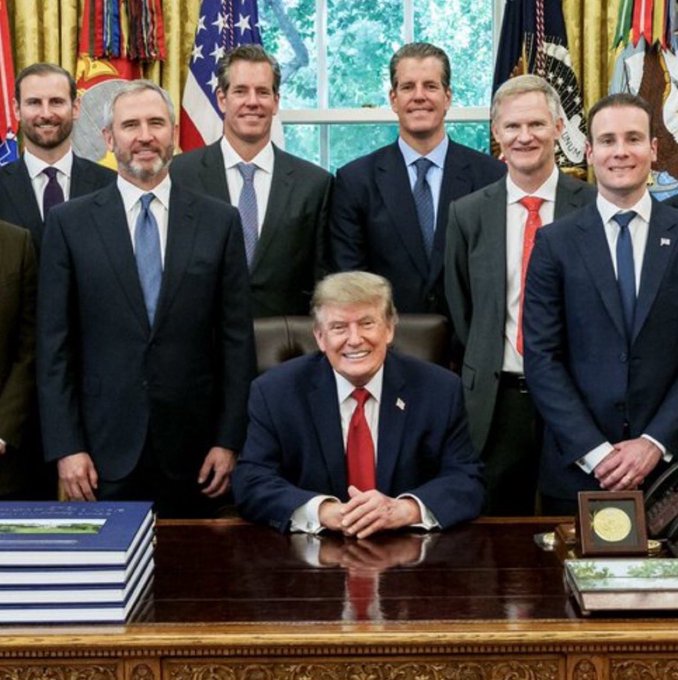 Donald Trump sits at the Resolute Desk in the Oval Office surrounded by a group of men in business suits, smiling for a group photo with American flags visible in the background.