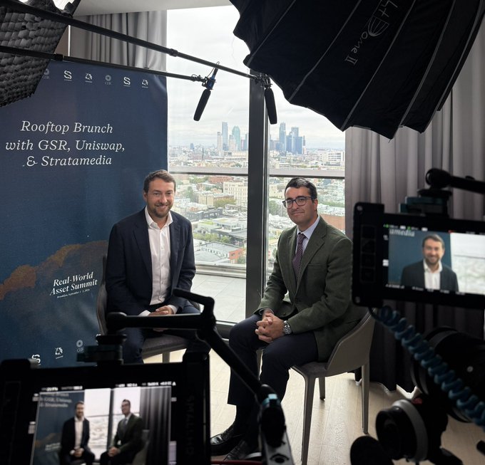 Two men in business attire sit for a video recording in front of a "Rooftop Brunch with GSR, Uniswap, & Stratamedia" backdrop, with a city skyline visible through the window, capturing a discussion fitting the post's theme of a big week for real-world assets (RWAs) and industry banter.