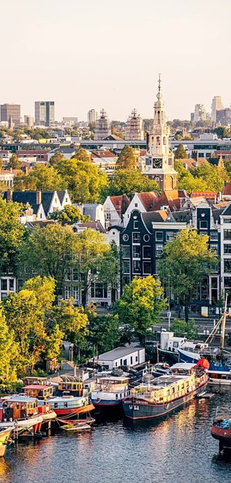 An aerial cityscape view of Amsterdam showing boats docked along a canal, historic buildings with stepped gables, and the Munttoren tower in the background.