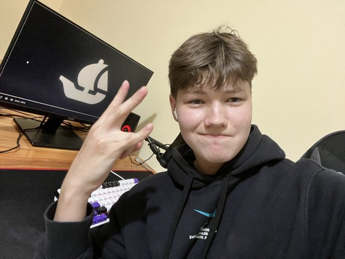 A smiling young man wearing a black hoodie throws up a peace sign while seated at a gaming setup featuring a monitor displaying the Opensea logo and a backlit keyboard, visually matching his post about an upcoming pixelated NFT collection dropping on Opensea on September 1st.