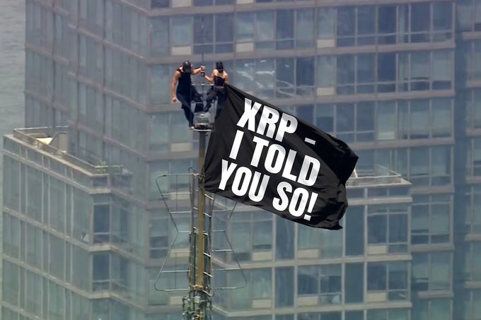 Two people unfurl a large black banner reading "XRP - I TOLD YOU SO!" from atop a skyscraper, parodying the iconic Spider-Man meme in a triumphant gesture toward those who doubted the XRP cryptocurrency.