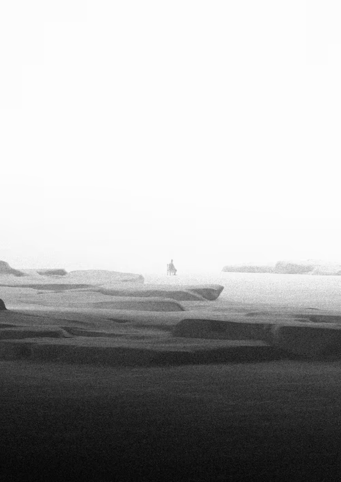 A hazy, high-contrast black and white photograph shows a tiny solitary figure standing on a rocky outcrop overlooking an expansive, mist-shrouded landscape.