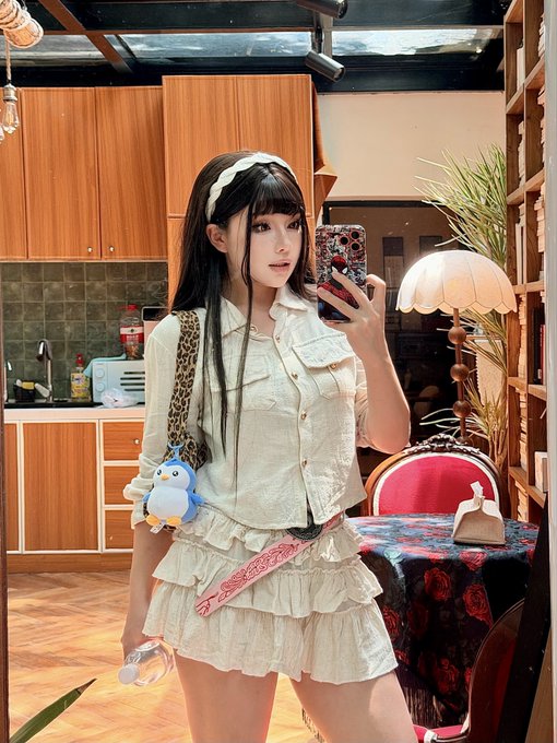 A young woman with long dark hair and bangs takes a mirror selfie while wearing a cream-colored blouse and matching ruffled skirt, accessorized with a white headband and a penguin plush charm on her bag strap. The cozy setting features warm wooden kitchen cabinets, a vintage-style table with a floral cloth, and a scalloped lamp, complementing the cheerful "Gmeow and Happy Saturday" vibe of her playful, cozy Saturday look.