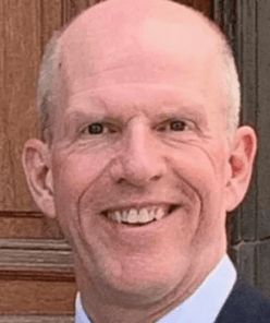 A professional headshot of a smiling, bald, middle-aged man in a suit, photographed against a wooden background.