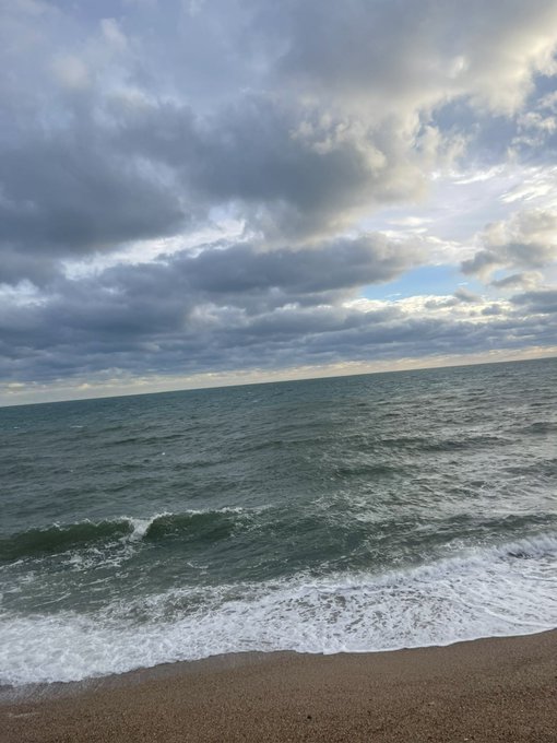 A serene coastal photograph showing ocean waves crashing onto a sandy beach under a dramatic cloudy sky with patches of blue.