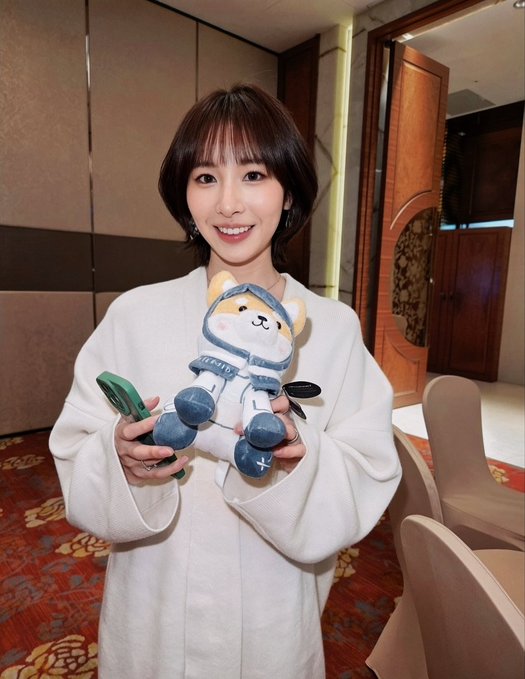 A smiling woman with short dark hair and a cream-colored sweater holds a plush dog toy dressed in an astronaut/spacesuit outfit while holding a phone.