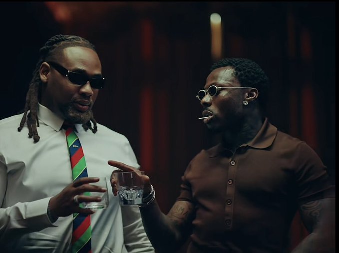 Two men at a formal gathering, one with dreadlocks wearing sunglasses, a white shirt, and a striped tie, the other in a brown polo shirt with round sunglasses, both holding glasses.