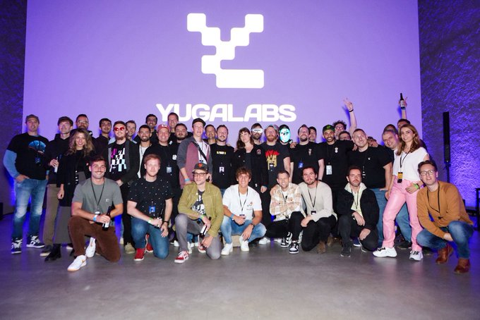 A large group of people poses for a team photo in front of a screen displaying the "YUGALABS" logo.