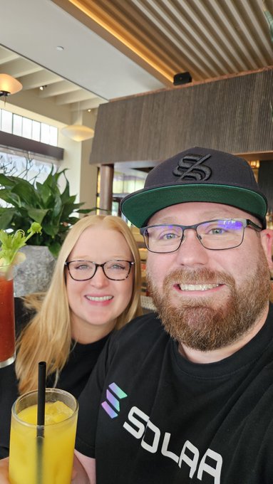 A couple takes a selfie at a restaurant; the man wears a black 'SOLAA' t-shirt and an 'SS' cap, while the woman smiles beside a glass of orange drink.