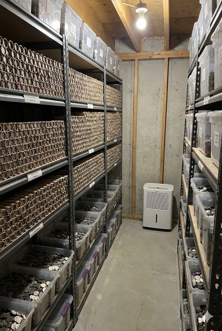 A storage room filled with shelves containing numerous cardboard coin tubes and plastic bins of coins, with a dehumidifier visible at the back.