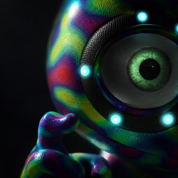 A 3D rendered close-up of a metallic, rainbow-iridescent spherical robot or creature with a large green eye and glowing cyan lights, reaching out with a small arm.