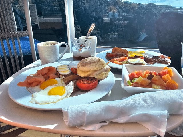 A breakfast spread on a round table features a plate with a fried egg, bacon, grilled tomato, pancakes, and potatoes, accompanied by a side of cut fruit and coffee, set against a backdrop of windows showing a scenic view.