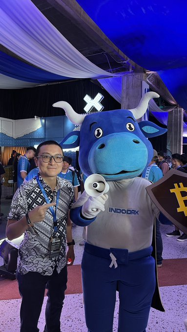 A smiling guest in glasses poses with his finger pointed up alongside the INDODAX blue bull mascot at the Coinfest Asia event, where the mascot displays the "INDODAX" logo on its shirt and a Bitcoin "₿" sign nearby. The scene reflects the post's invitation to visitors stopping by the INDODAX booth, where attendees can try out features like the "Freedom Decibel."