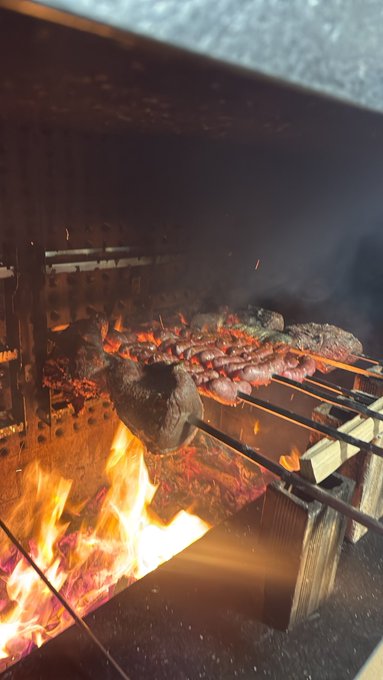 Meat roasts over an open wood fire inside a metal grill, with flames, glowing coals, smoke, and sparks visible.