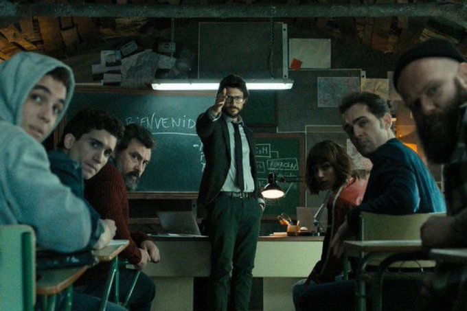 A group of people sit in a dim classroom with a chalkboard reading "BIENVENID" while a man in a suit and glasses points forward, appearing to be from a Spanish thriller series.