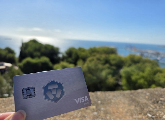A hand holds a metal Visa debit card from Crypto.com, featuring the company's hexagonal logo, against a scenic backdrop of lush green trees and ocean. The card ties into the post's message about earning $CRO cashback rewards, showcasing the physical card used to unlock crypto cashback on everyday purchases.