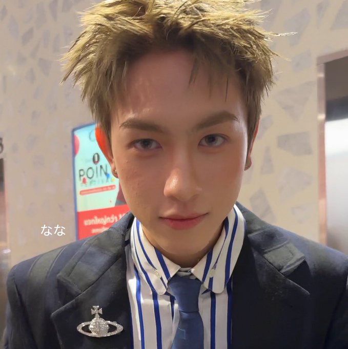 A young man with spiky blonde hair, wearing a navy blazer over a blue-and-white striped shirt and blue tie, looks directly at the camera with a solemn expression, his sorrowful demeanor reflecting the post's announcement that an auntie has passed away during his time at the Chidchom event with PROXIE members Onglee and Th.