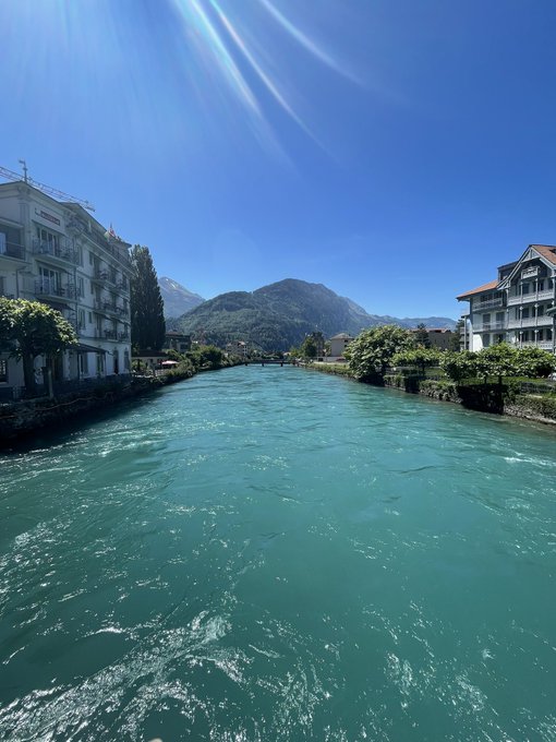 The image shows a beautiful view of a river flowing through a town, surrounded by buildings. The sky above the water appears blue with some clouds scattered across it. In the background, there are mountains visible, adding to the picturesque scenery. The photo is taken from a boat on the river, providing an interesting perspective of the landscape below.