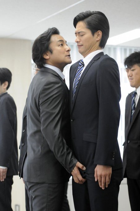 Two Asian men in black business suits stand face-to-face in what appears to be an office setting, with the man on the left looking up at the much taller man on the right who wears a striped blue and white tie. Other suited men are visible in the background, observing the intense exchange.