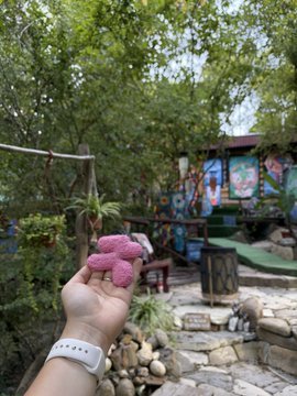 A hand holding pink frosted heart-shaped cookies in front of an outdoor garden setting with greenery, colorful murals, and stone seating.