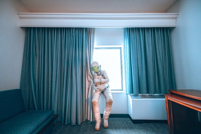 The image shows a man dressed as an astronaut standing near a window, wearing a full spacesuit that includes boots, a helmet, and holding onto a railing with his right hand while leaning against the curtain of the room. There are also blue curtains visible in the background, which match the color of his suit. The scene appears to be part of a promotional or artistic project centered around space exploration.