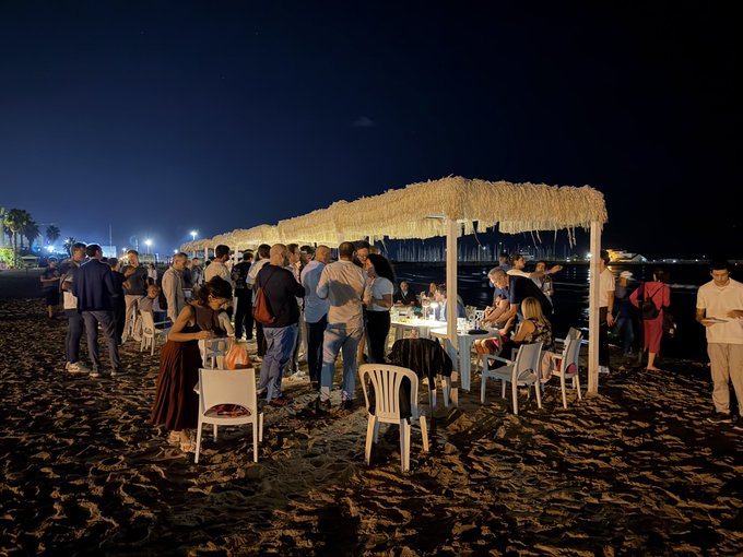 A nighttime beach gathering with people socializing under a thatched-roof structure, with chairs and a table set up on the sand near the water.