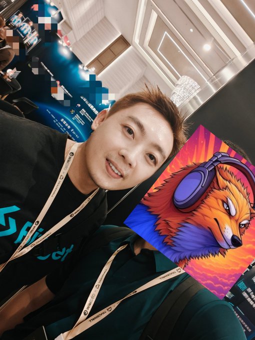 A young man takes a selfie with a colorful card depicting an orange bear character wearing purple headphones, wearing a lanyard with 'THRESHOLD' text.