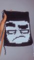 A small black handcrafted pouch or coin purse featuring an embroidered deadpan/sad face design, held against a light background.