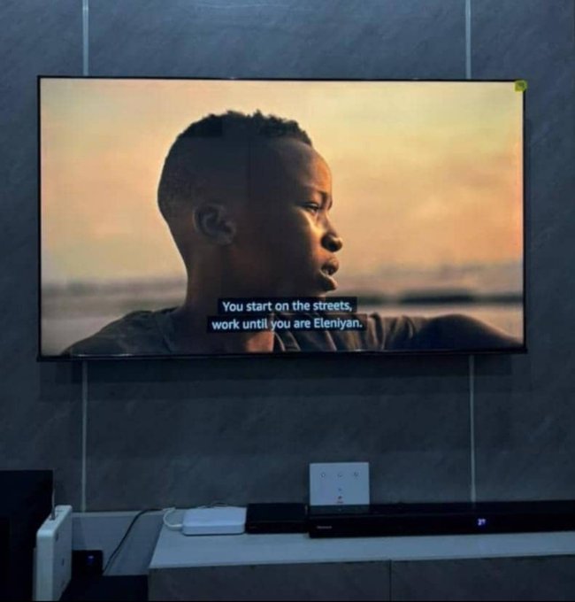 A wall-mounted TV displaying a movie scene with subtitles reading 'You start on the streets, work until you are Eleniyan,' showing a man's profile against a sunset background.