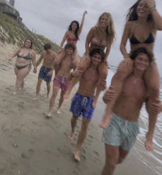 A group of young adults in swimwear play on a sandy beach, with women sitting on the men's shoulders as they walk along the shoreline.
