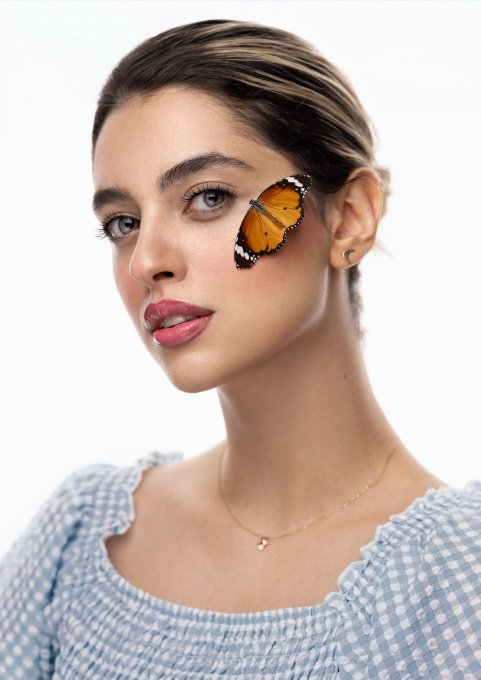 A close-up portrait of a young woman with a monarch butterfly resting on her cheek, wearing a blue and white gingham top and a delicate gold necklace.